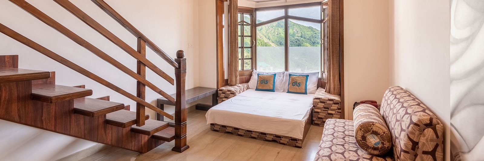 A spacious room near Bhagsunag Temple featuring a wooden staircase, a bed, a sofa, and a large window offering a mountain view – DLS Meghavan Resort.